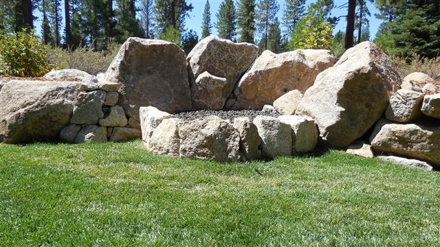 Granite boulder fire pit set into a mown lawn against a boulder retaining wall